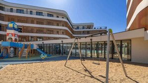 Un parque infantil con tobogán y columpios, una zona de arena y un edificio moderno con balcones y ventanas amplias.
