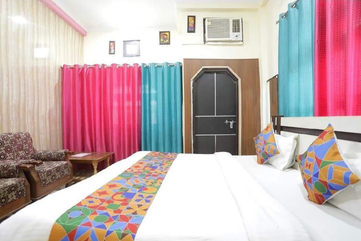 A hotel room with a bed, a chair, a small table, curtains, a door, and an air conditioning unit on the wall.