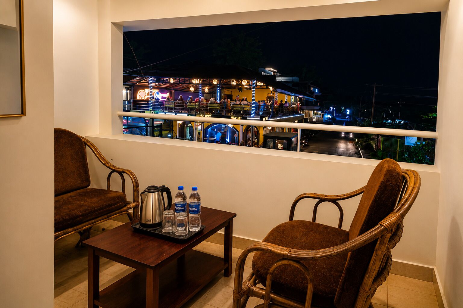 Un balcon avec deux fauteuils en osier, une petite table avec une bouilloire et des bouteilles d’eau, et une vue sur un immeuble illuminé la nuit.