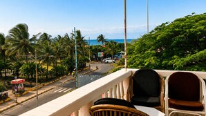 A balcony with wicker chairs and a table, overlooking a street with palm trees and a view of the ocean. - Holiday Beach Resort (Candolim)