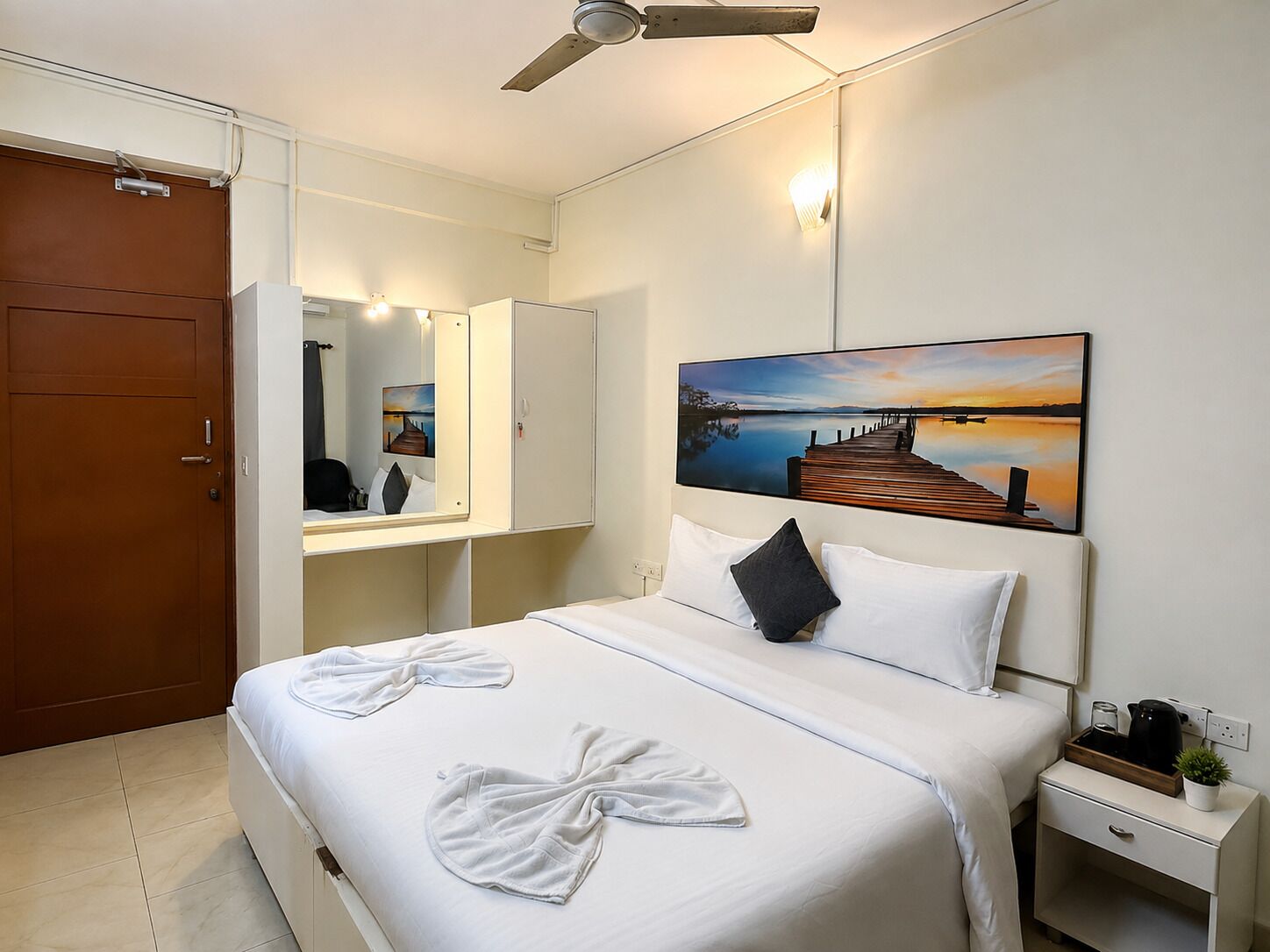 Una habitación de hotel con cama, mesita de noche, cuadro, ventilador de techo y puerta.