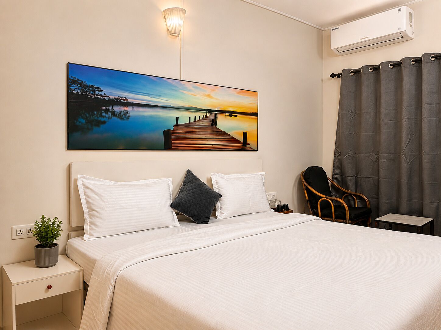 Une chambre d’hôtel avec un lit, une table de chevet ornée d’une plante, une chaise, un tableau représentant un coucher de soleil sur l’eau et un appareil de climatisation.