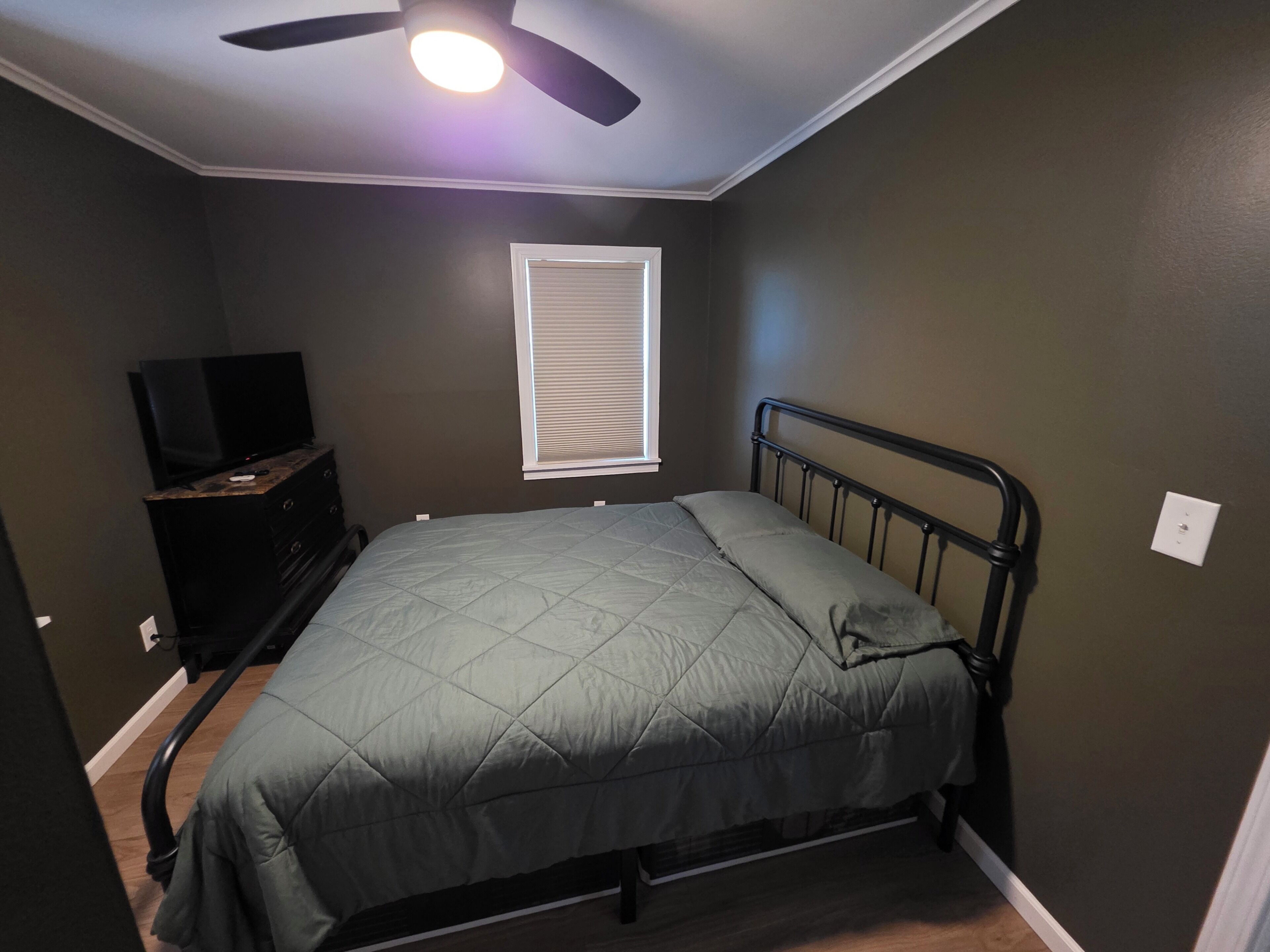 Een slaapkamer met een bed, een plafondventilator, een raam met zonwering en een dressoir met een televisie.