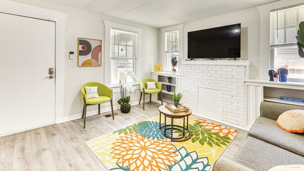 Een woonkamer met een wit bakstenen haard, een flatscreen televisie erboven en een kleurrijk vloerkleed. Er staan twee groene stoelen met witte kussens en een kleine ronde tafel met een plant.