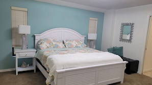 Une chambre à coucher avec un lit blanc, deux tables de chevet, une commode, un miroir et un sol recouvert d’une moquette.