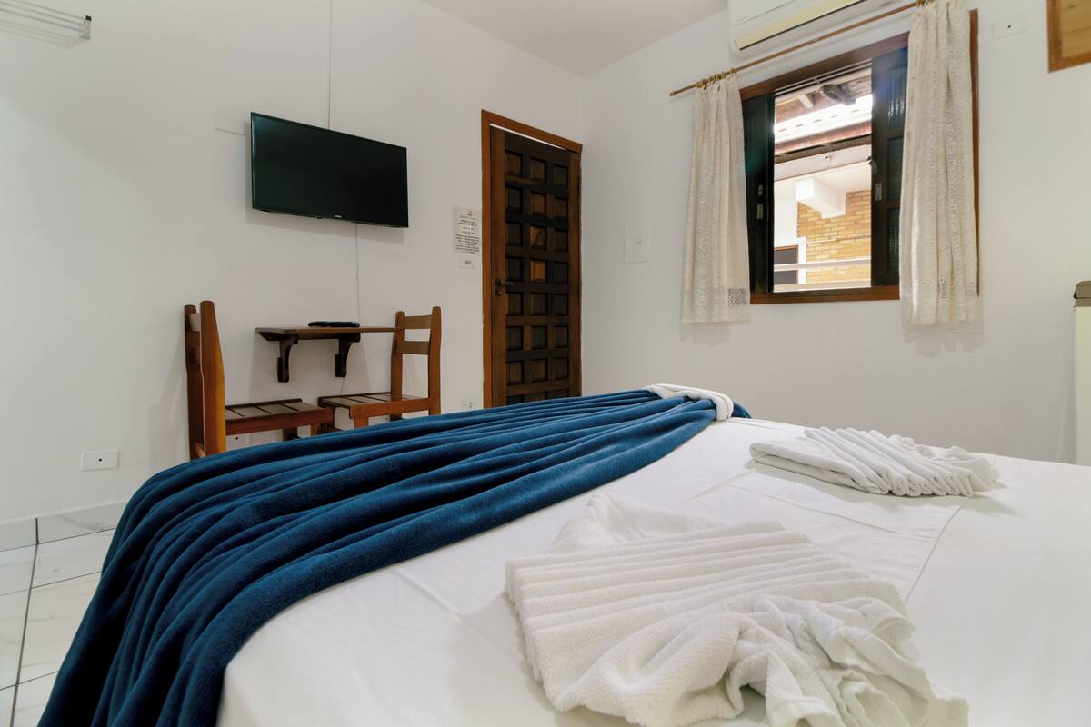 A bedroom with a bed, a TV mounted on the wall, a wooden door, a window with curtains, and a small table with a chair.