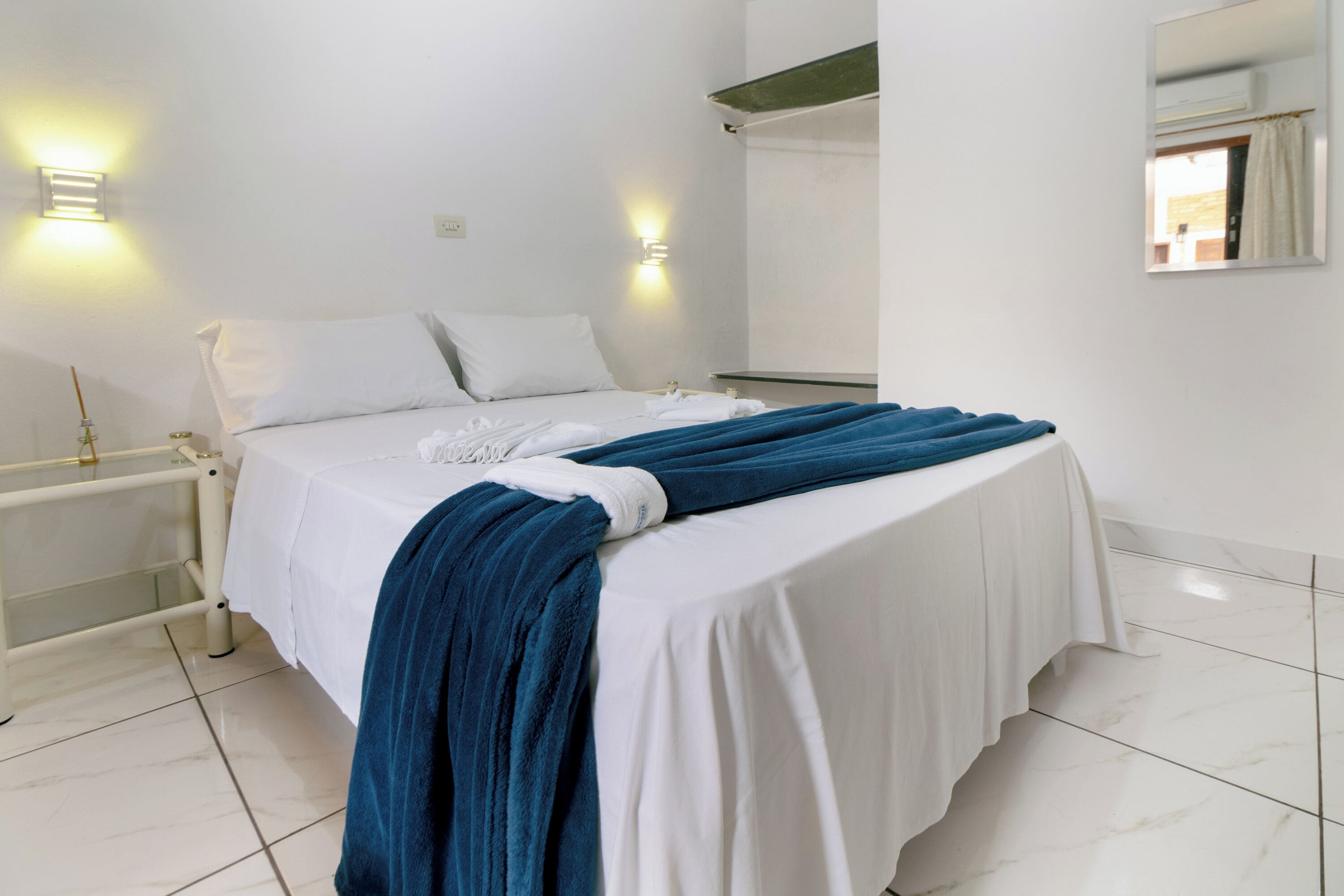 Una habitación de hotel con cama, paredes blancas, dos mesitas de noche y un espejo.