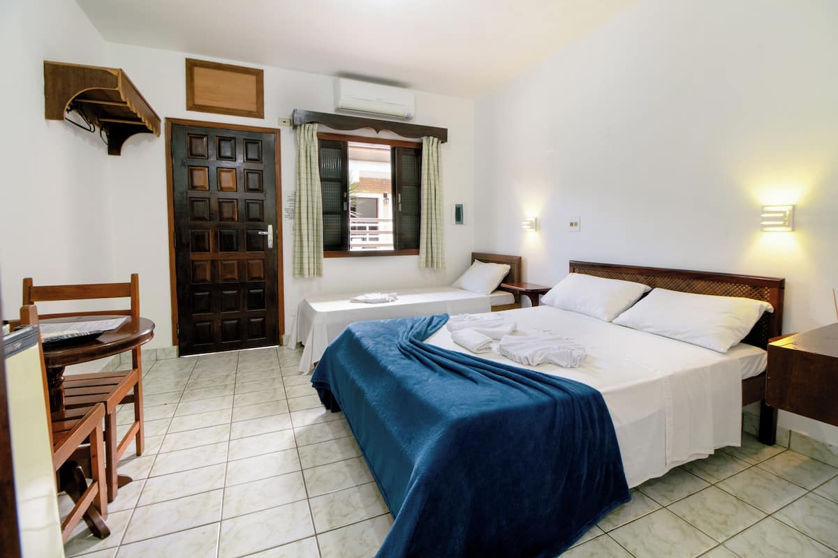 A bedroom with a bed, a desk, and a chair. There is a window with curtains, a wall-mounted air conditioner, and a wall-mounted light fixture.
