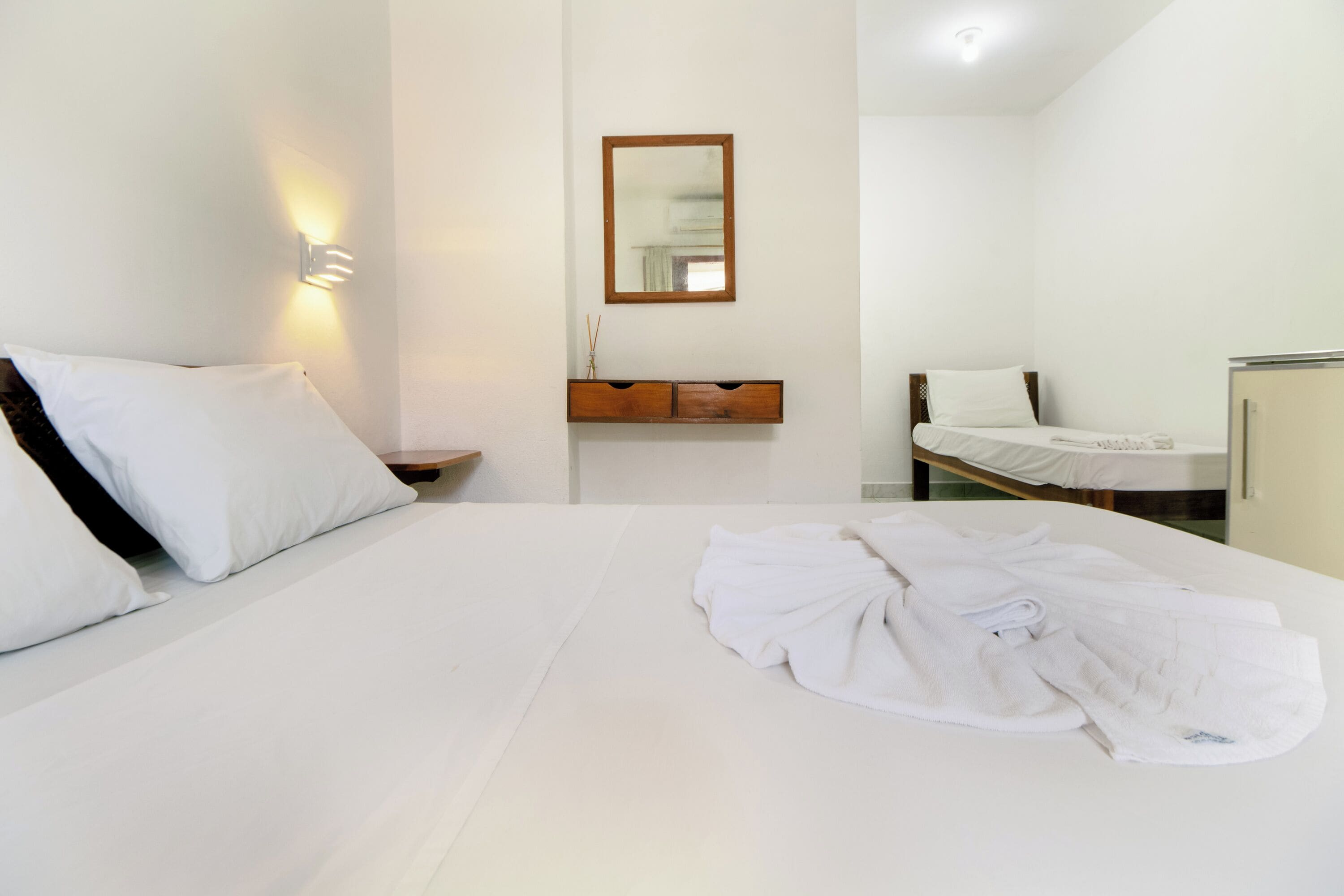 Quarto de hotel com uma cama grande, uma mesinha de cabeceira, um espelho e uma pequena mesinha com uma luminária.