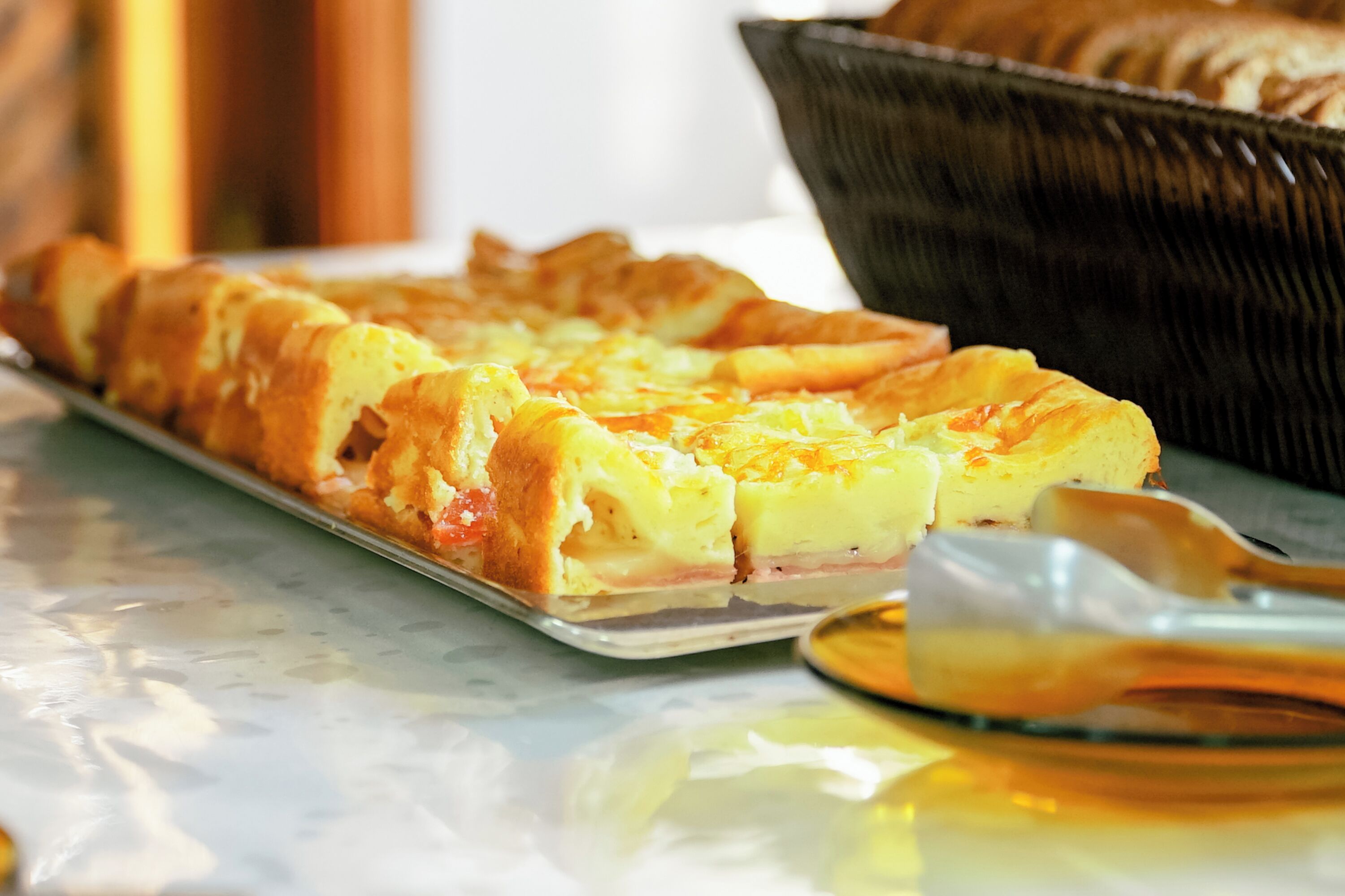 Una bandeja de quiche con jamón y queso, una cesta de pan y una cuchara sobre una superficie de mármol.