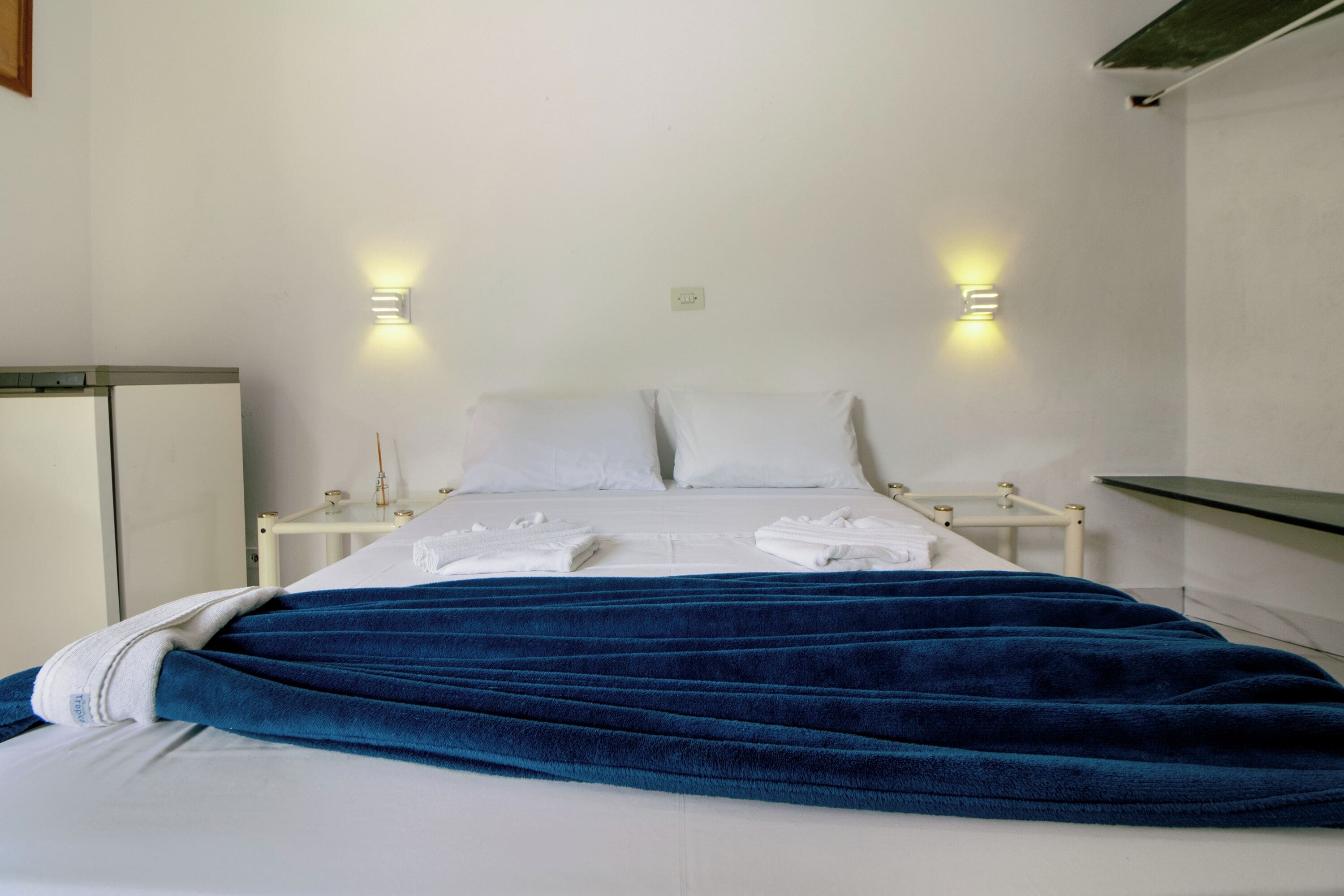 Una cama bien hecha con una colcha azul, almohadas blancas y dos lámparas de noche.
