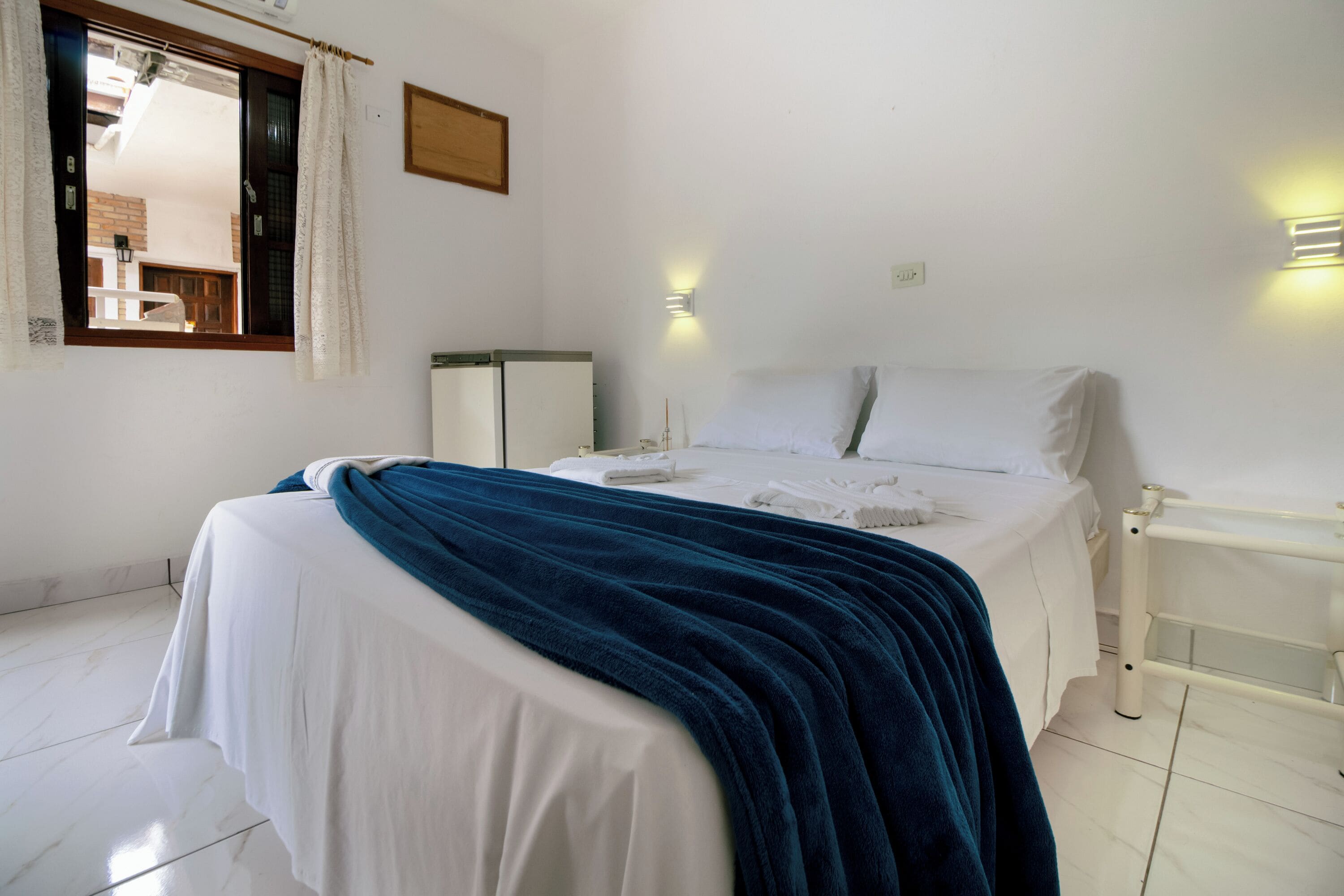 A hotel room with a bed, a bedside table, a nightstand, a window with curtains, and a wall-mounted light fixture.