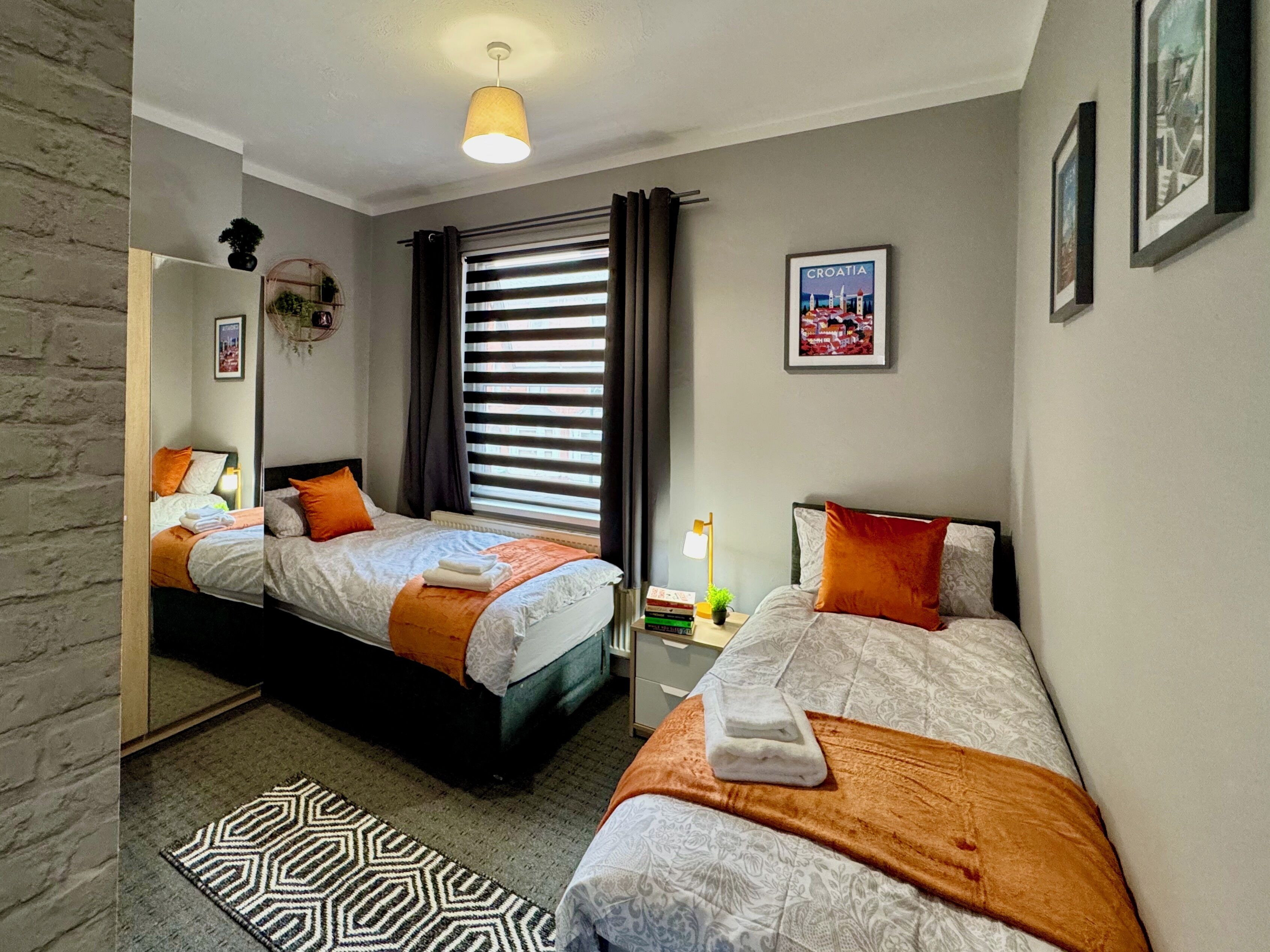 Una habitación de hotel con dos camas, un ventanal con cortinas, una mesita con lámpara y un cuadro en la pared.