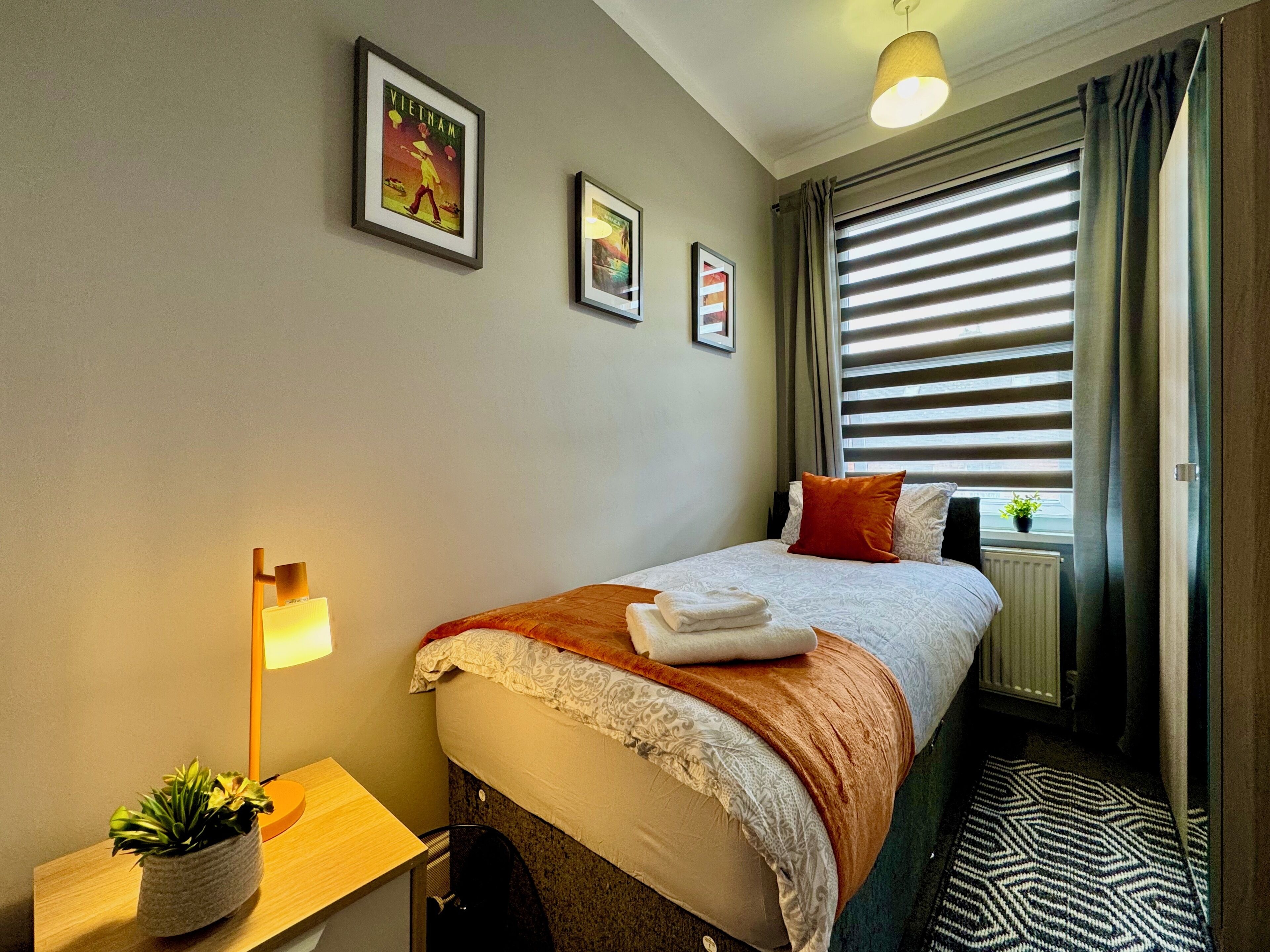 Une chambre d’hôtel bien rangée, avec un lit, une table de chevet dotée d’une lampe et une fenêtre avec des stores.