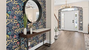 A hallway with a floral wallpaper, a round mirror, a wooden console table, and a potted plant. - Hilltop 6BR Disney Villa: Theater, Pool/Spa, View (Kissimmee)