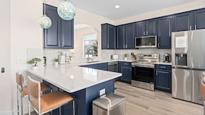 A modern kitchen with dark blue cabinets, stainless steel appliances, a large island with bar stools, and pendant lighting. - Hilltop 6BR Disney Villa: Theater, Pool/Spa, View (Kissimmee)