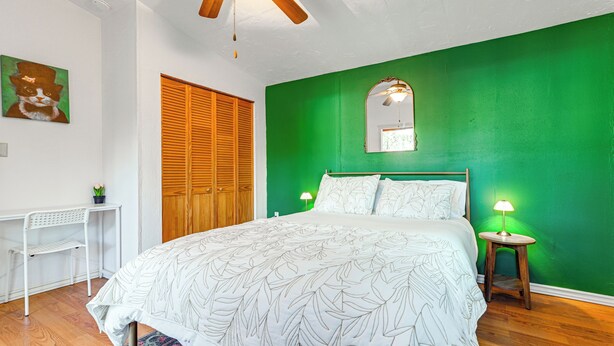 A bedroom with a green wall, a bed with a leaf-patterned bedspread, a wooden ceiling fan, a white desk with a plant, and a wooden door. - Grants Pass Cottage ~ 8 Mi to Rogue River! (Grants Pass)