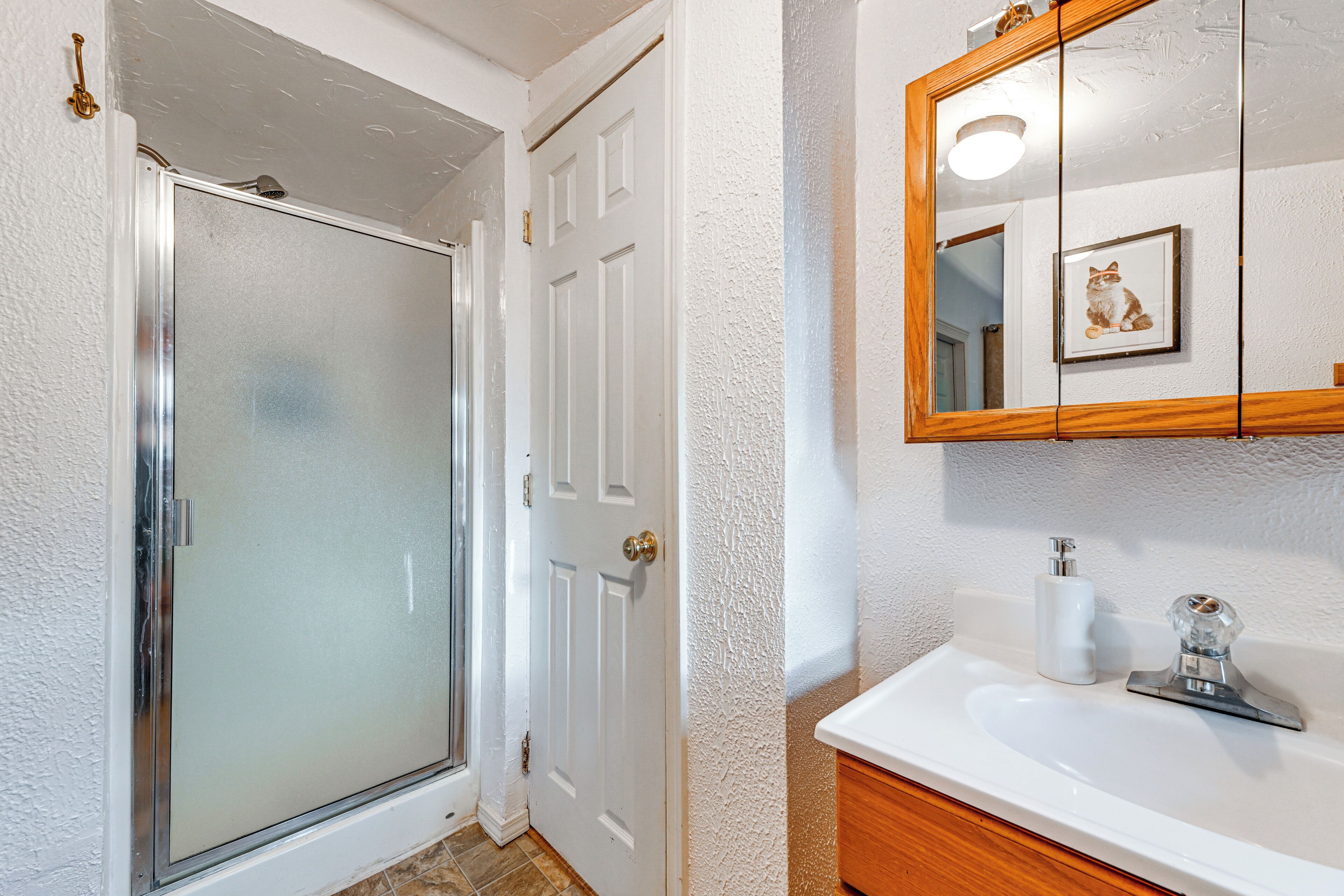 A bathroom with a glass shower door, a white sink with a wooden vanity, and a mirror with a framed picture.