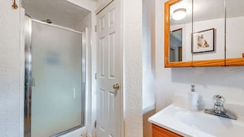 A bathroom with a glass shower door, a white sink with a wooden vanity, and a mirror with a framed picture.