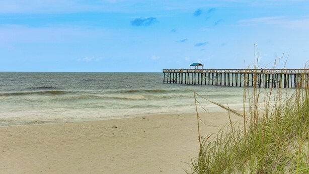 A beach with a wooden pier extending into the ocean, a small structure on the pier, and sand in the foreground.