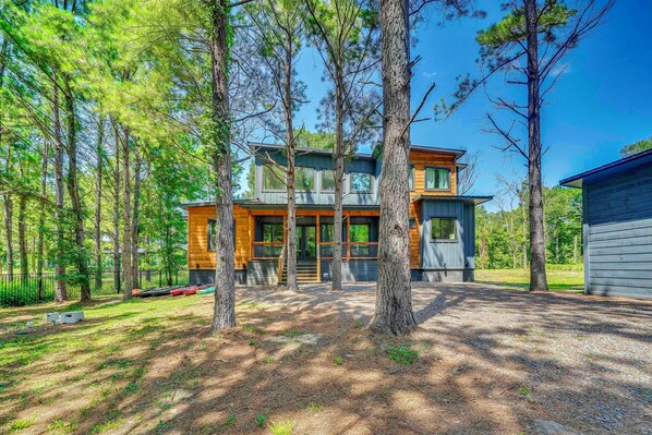 A modern two-story cabin with a large deck area, surrounded by a forested landscape.