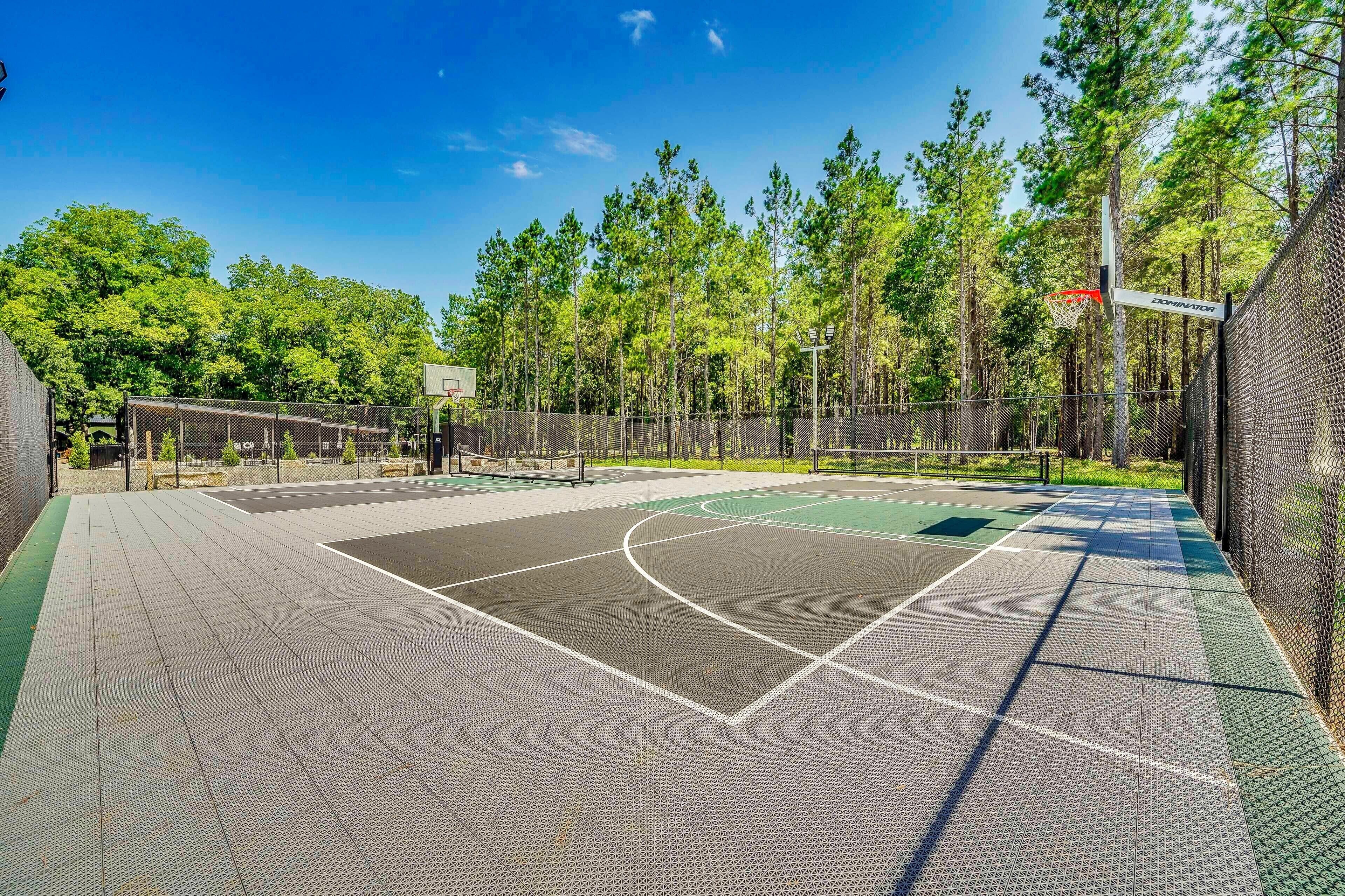 An outdoor basketball court with a chain-link fence, trees, and a clear sky.