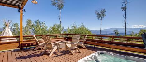 Um deck de madeira com mobiliário para área externa e uma banheira de hidromassagem, com vista para uma paisagem deslumbrante com árvores e montanhas.