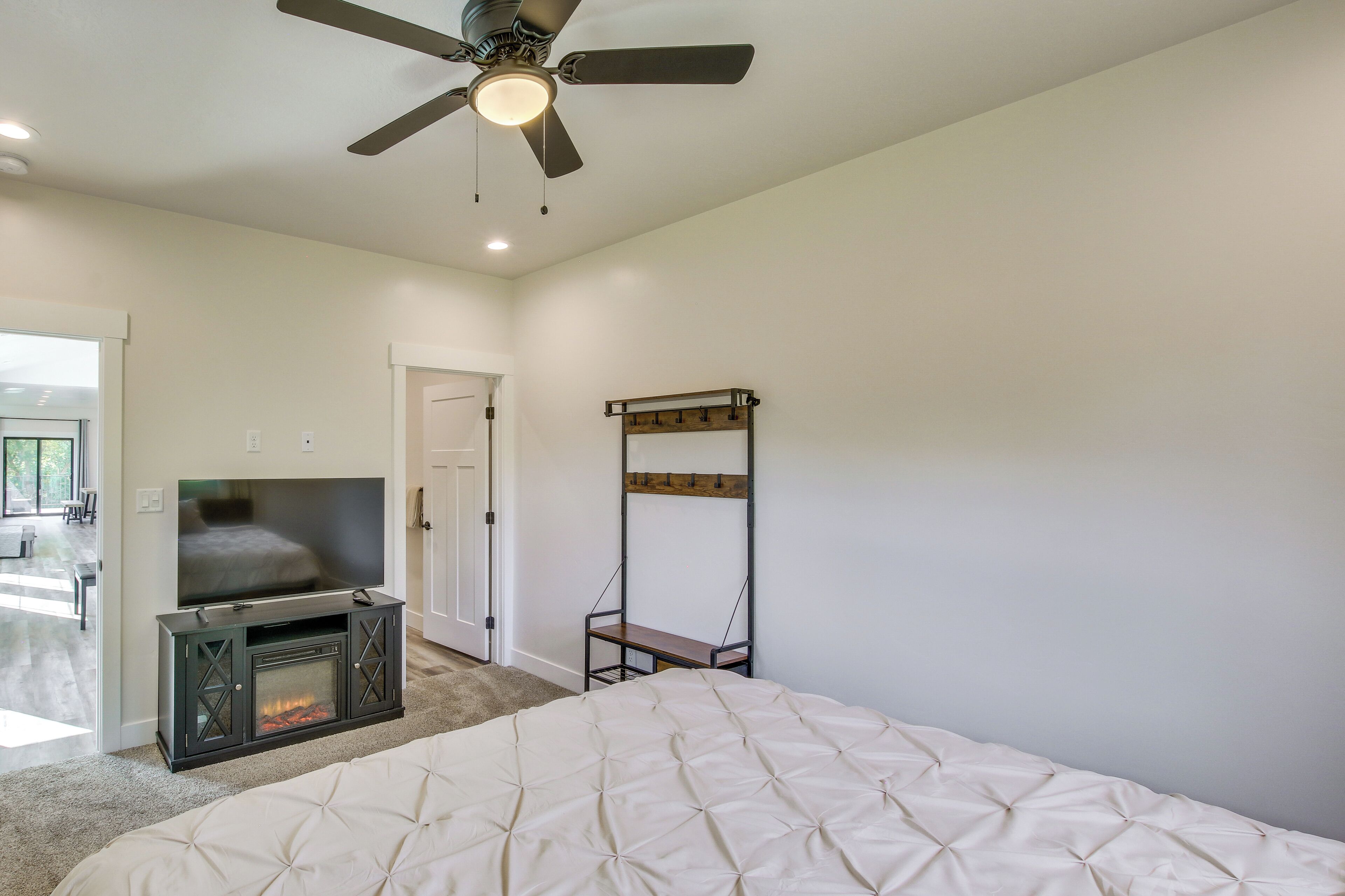 A bedroom with a bed, a TV, a fireplace, a ceiling fan, and a door leading to another room.