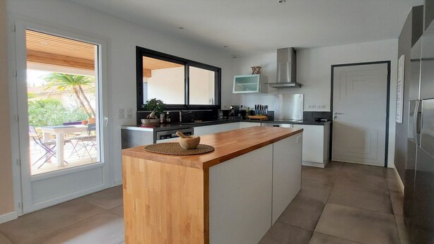 A modern kitchen with a wooden island, stainless steel appliances, and a large window overlooking a patio area. - Villa Plain Pied (Fayence)