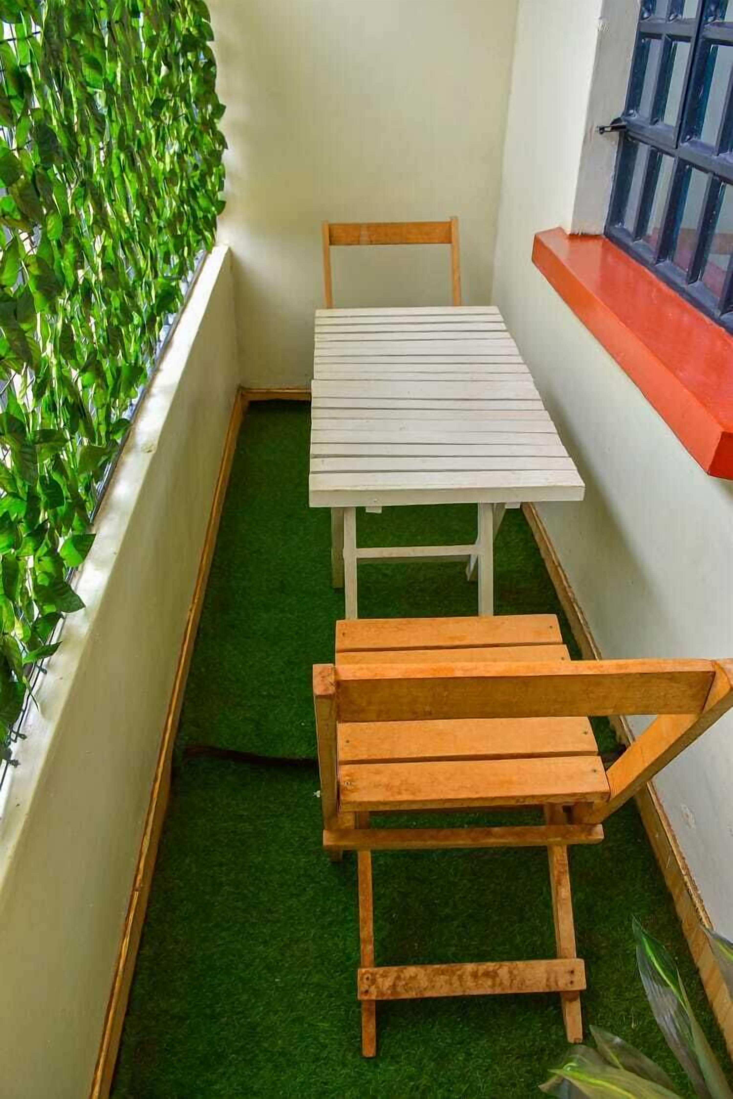 A small room with a white table and two wooden chairs, a green artificial grass floor, and a wall with green foliage.