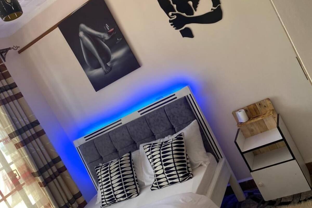 A bedroom with a bed, a headboard with blue lighting, a wall with a silhouette artwork, a poster, and a small wooden nightstand.