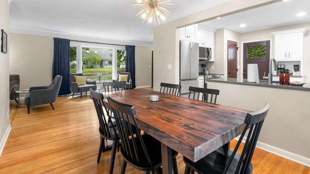A modern dining area with a wooden table and chairs, a kitchen with white cabinets, and a view of the outdoors.