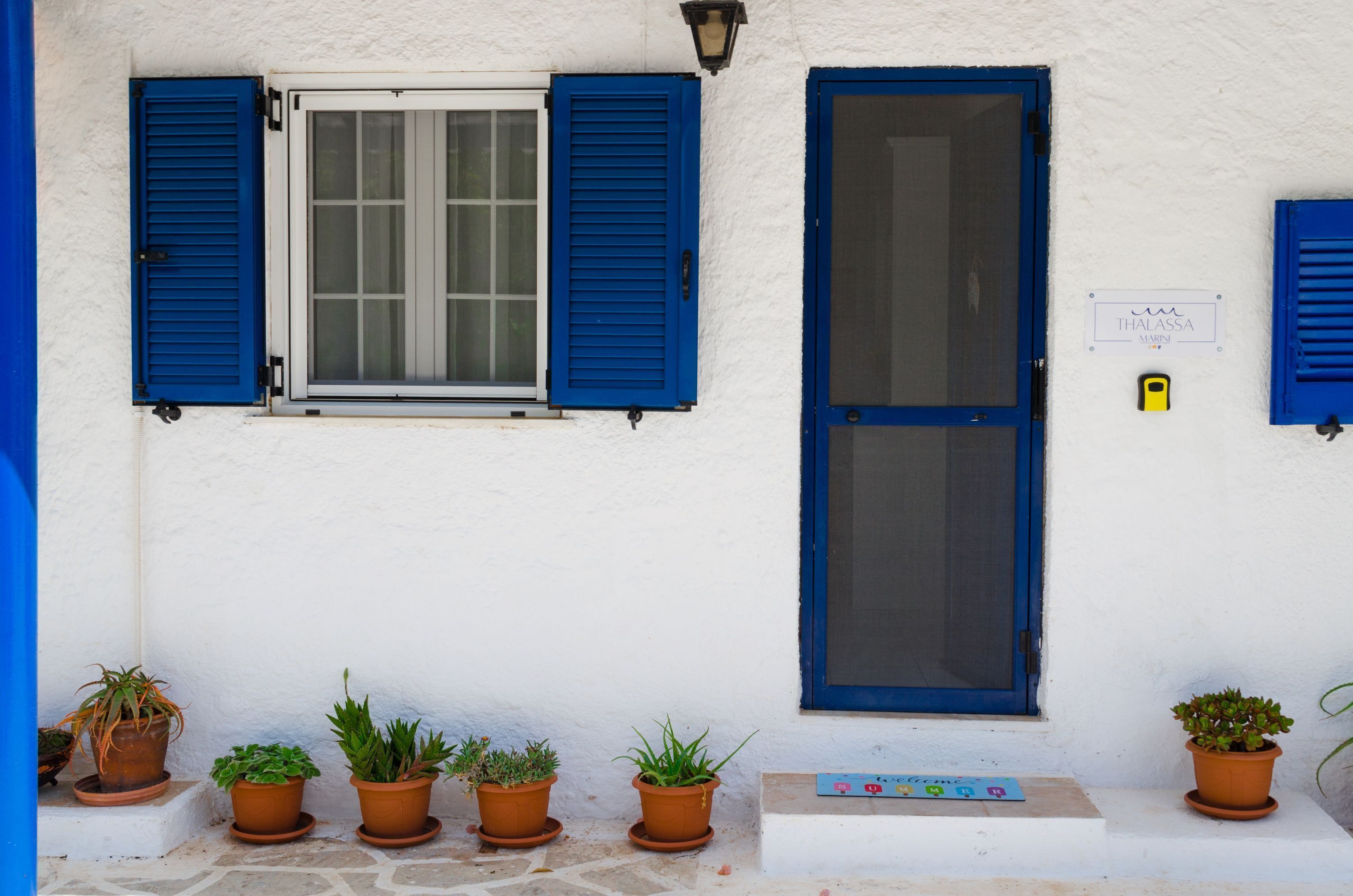 Facciata di un edificio con parete bianca, una porta blu e una finestra con persiane azzurre. Vasi con piante davanti alla porta.