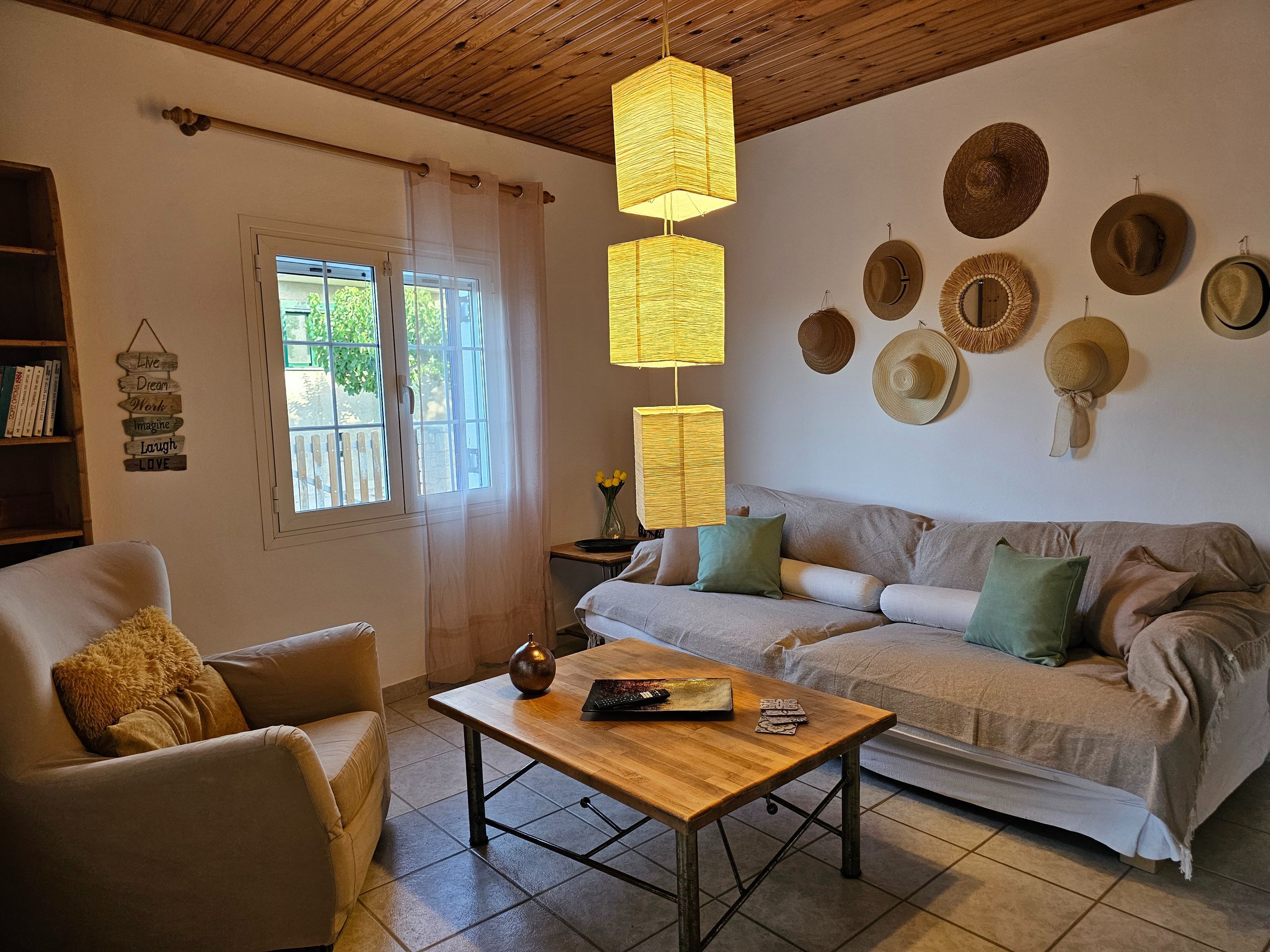 A cozy living room with a sofa, armchair, and a coffee table. There are decorative hats on the wall and a window with curtains.