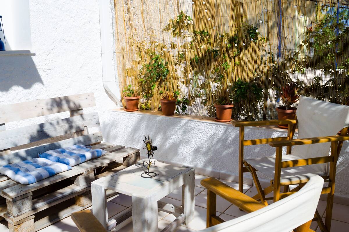 A wooden outdoor seating area with a bench and a small table, surrounded by potted plants and a white wall.