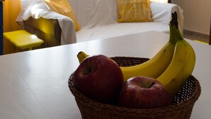 A room with a white sofa, yellow curtains, a basket of fruit on a table, and a window with natural light. - Marini Traditional Cottages (Aegina)