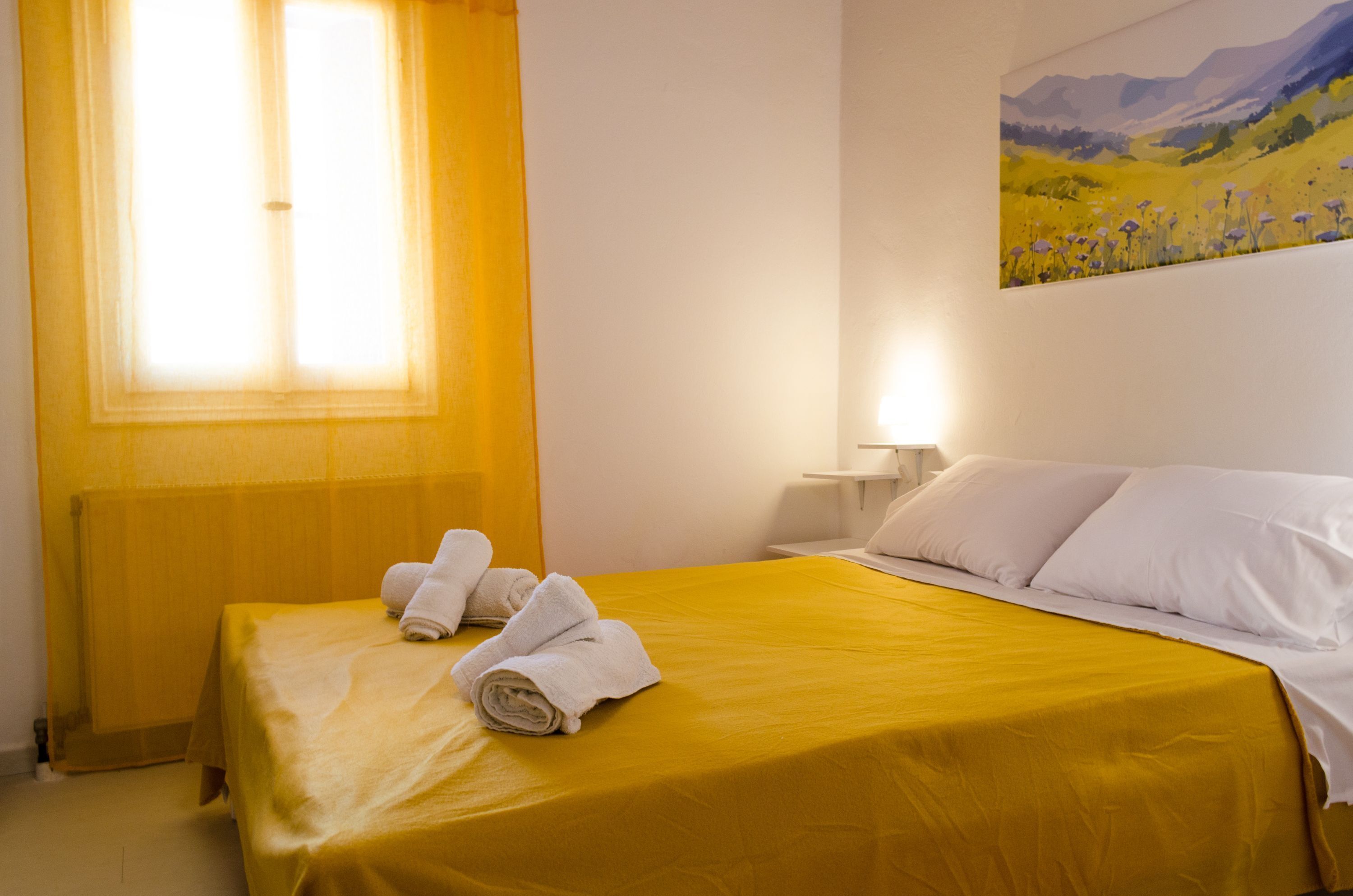 Una habitació d'hotel amb un estovat groc, almoines blanques, un quadre a la paret i una finestra amb cortines grogues.