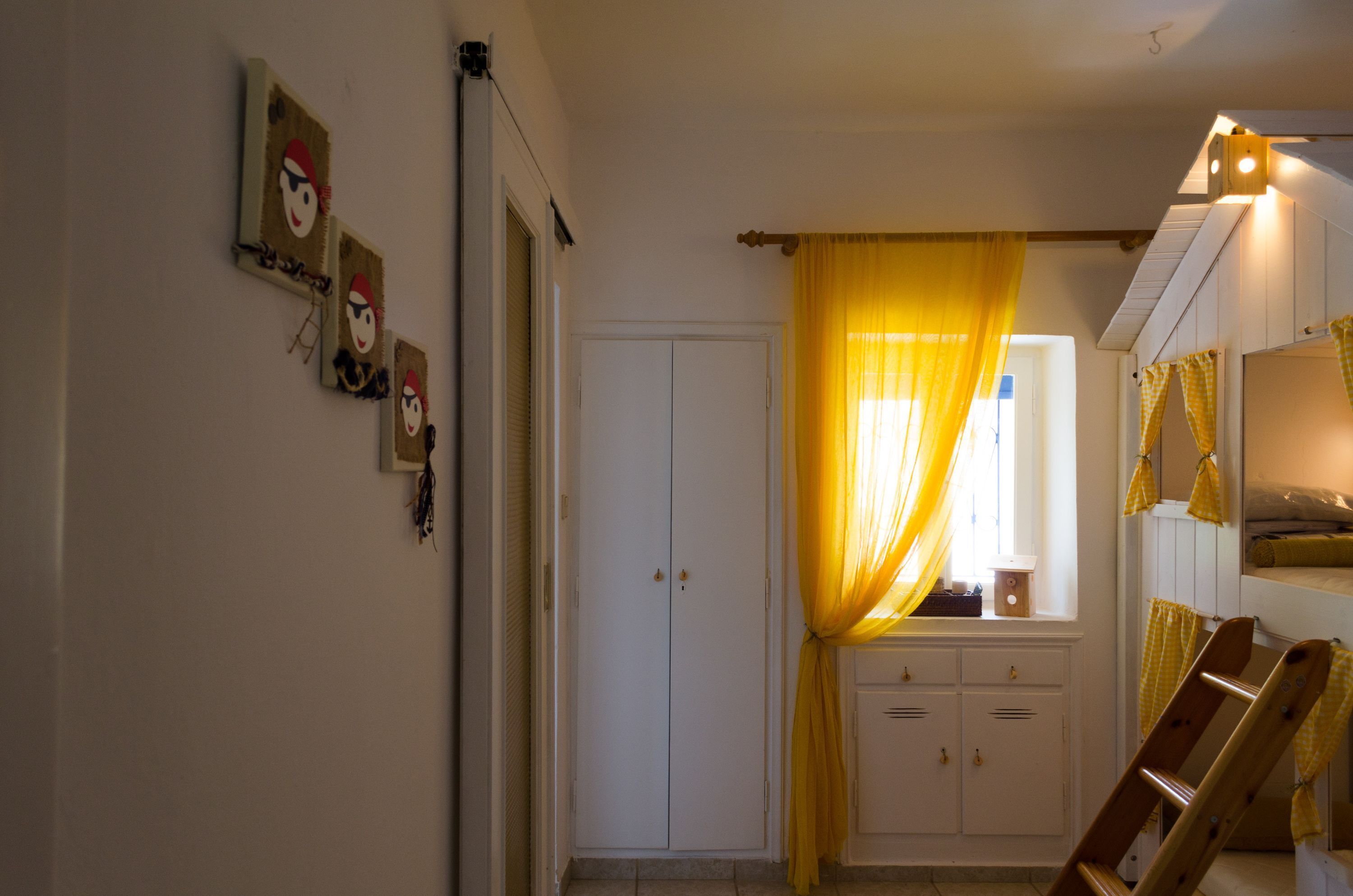 A room with a wardrobe, a window with yellow curtains, a ladder, and wall-mounted artwork.