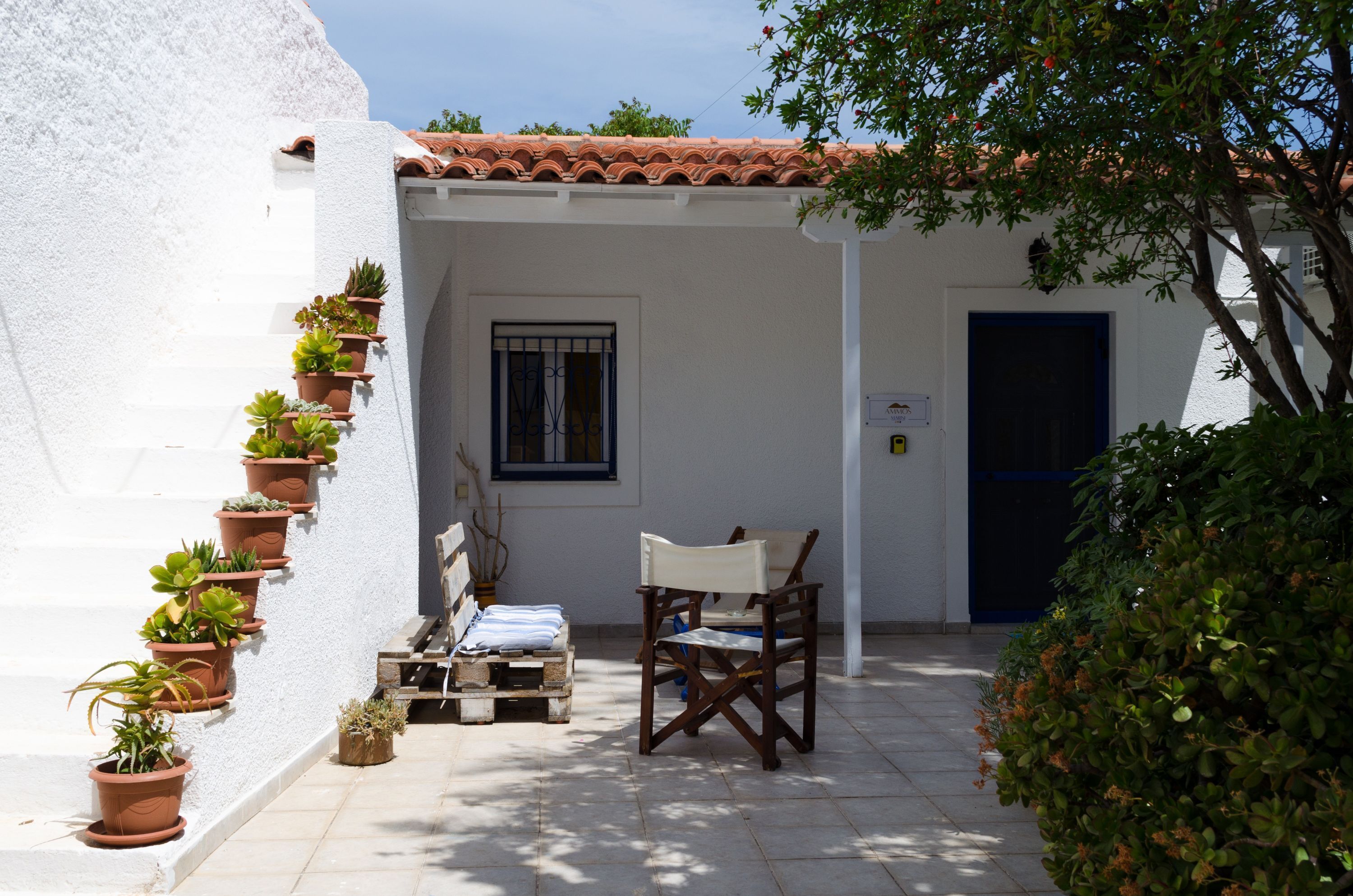 A white building with terracotta roof tiles, a wooden chair, and potted plants on the steps.