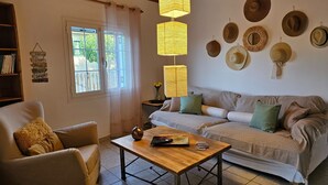 A cozy living room with a sofa, armchair, and coffee table, a bookshelf, and a window with curtains. - Marini Traditional Cottages (Aegina)