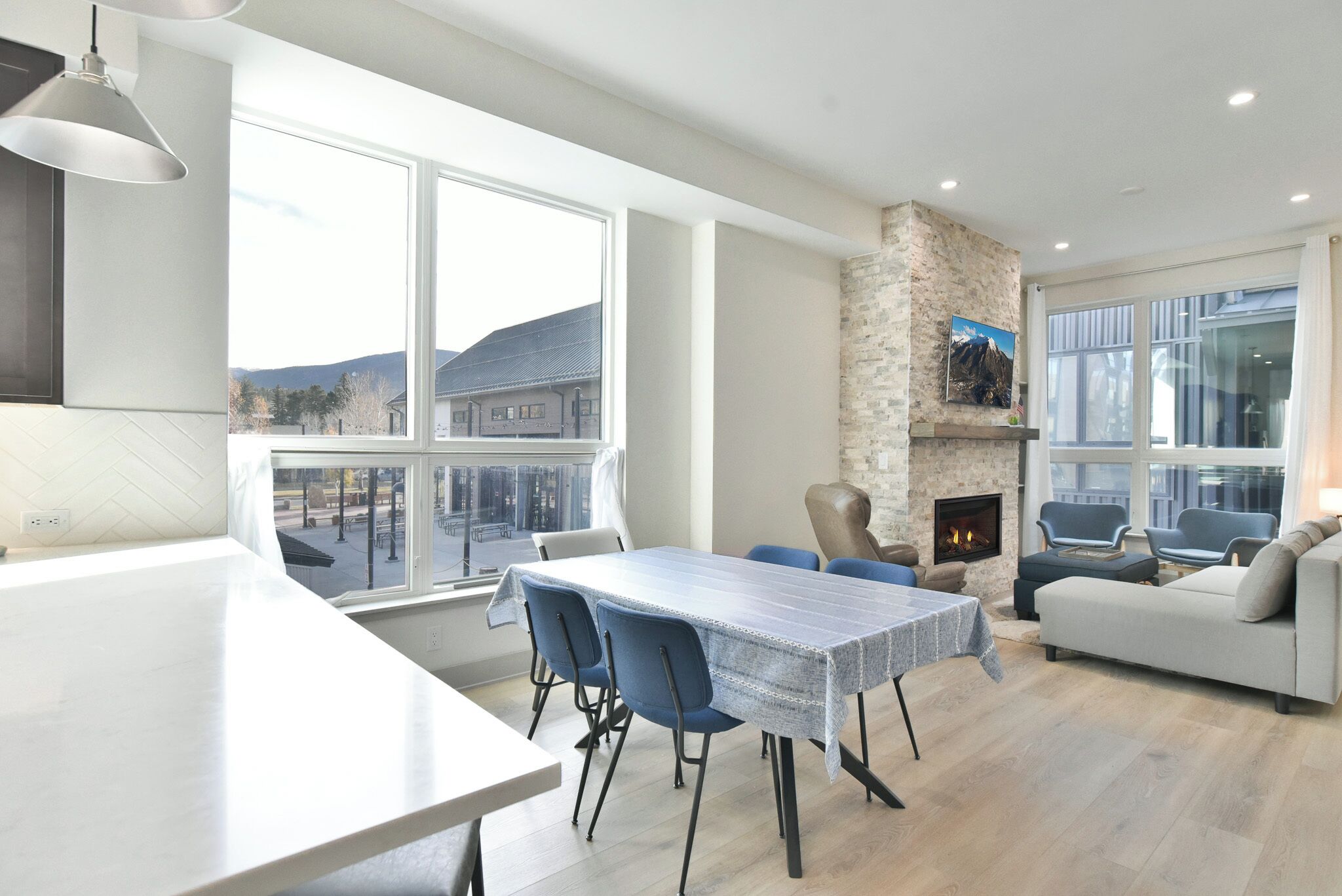 A modern living room with a fireplace, a dining area with a table and chairs, and large windows offering a view of the mountains.
