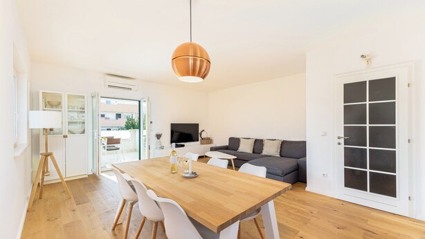 A modern living space with a wooden dining table, white chairs, a grey sofa, and a large window. - Olive Leaf Kastela - First Floor Apartment (Kastela)