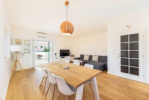 A modern living space with a wooden dining table, white chairs, a grey sofa, and a large window.