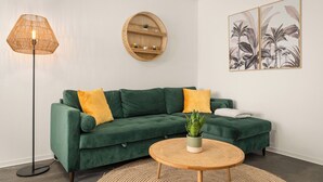 A green sofa with yellow cushions, a round table with a plant, a floor lamp, and a wall-mounted shelf with decorative items. - Kamingo for 12 people - Central, terrace, PS4 & Stream (Frankenberg (Eder))