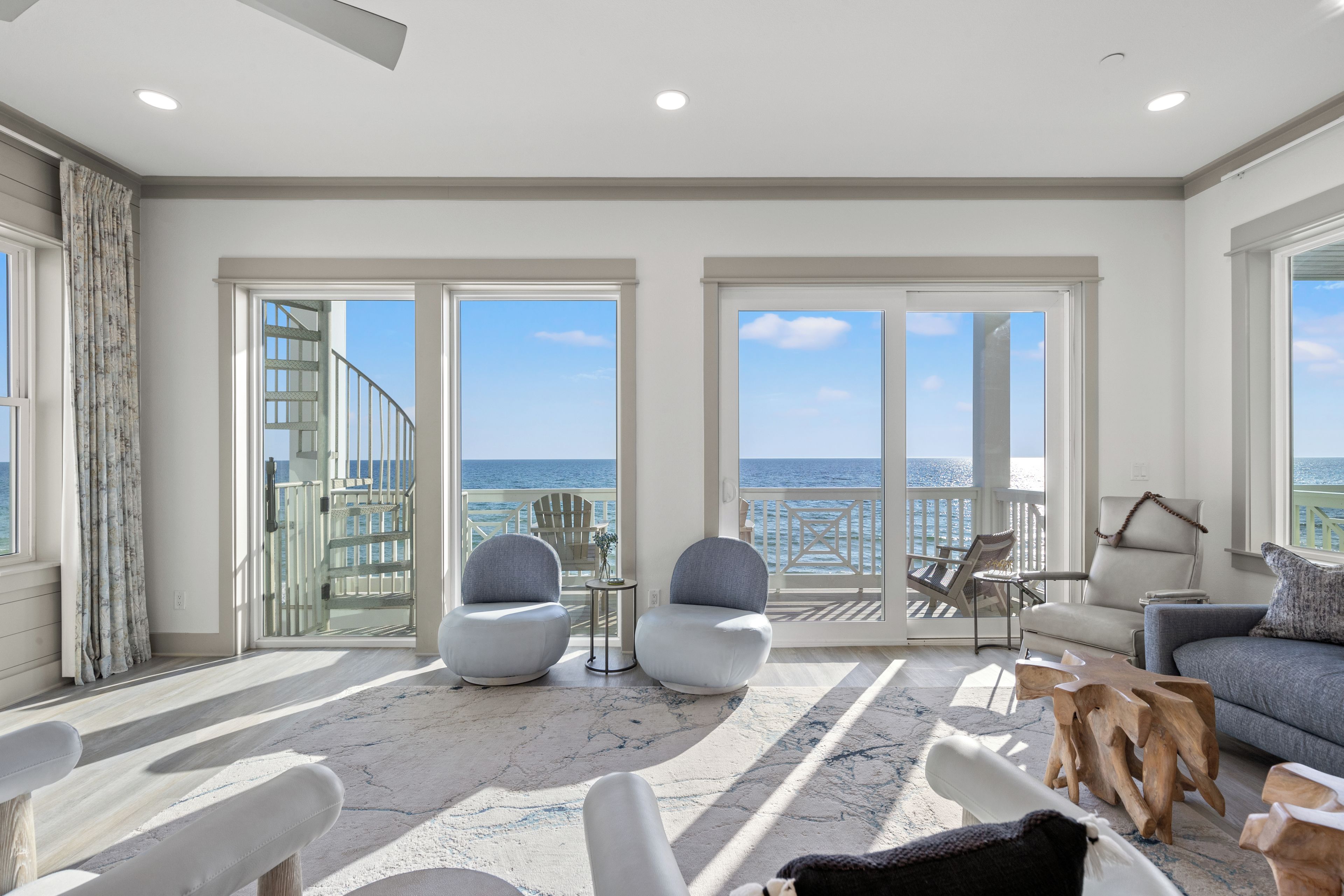 Een ruime kamer met een grote schuifdeur die uitkomt op een balkon met uitzicht op zee. Er staan twee fauteuils, een bank en een kleine tafel met een lamp.