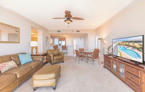 A spacious living room with a brown leather sofa, a dining area with a table and chairs, a television on a wooden cabinet, and a ceiling fan.