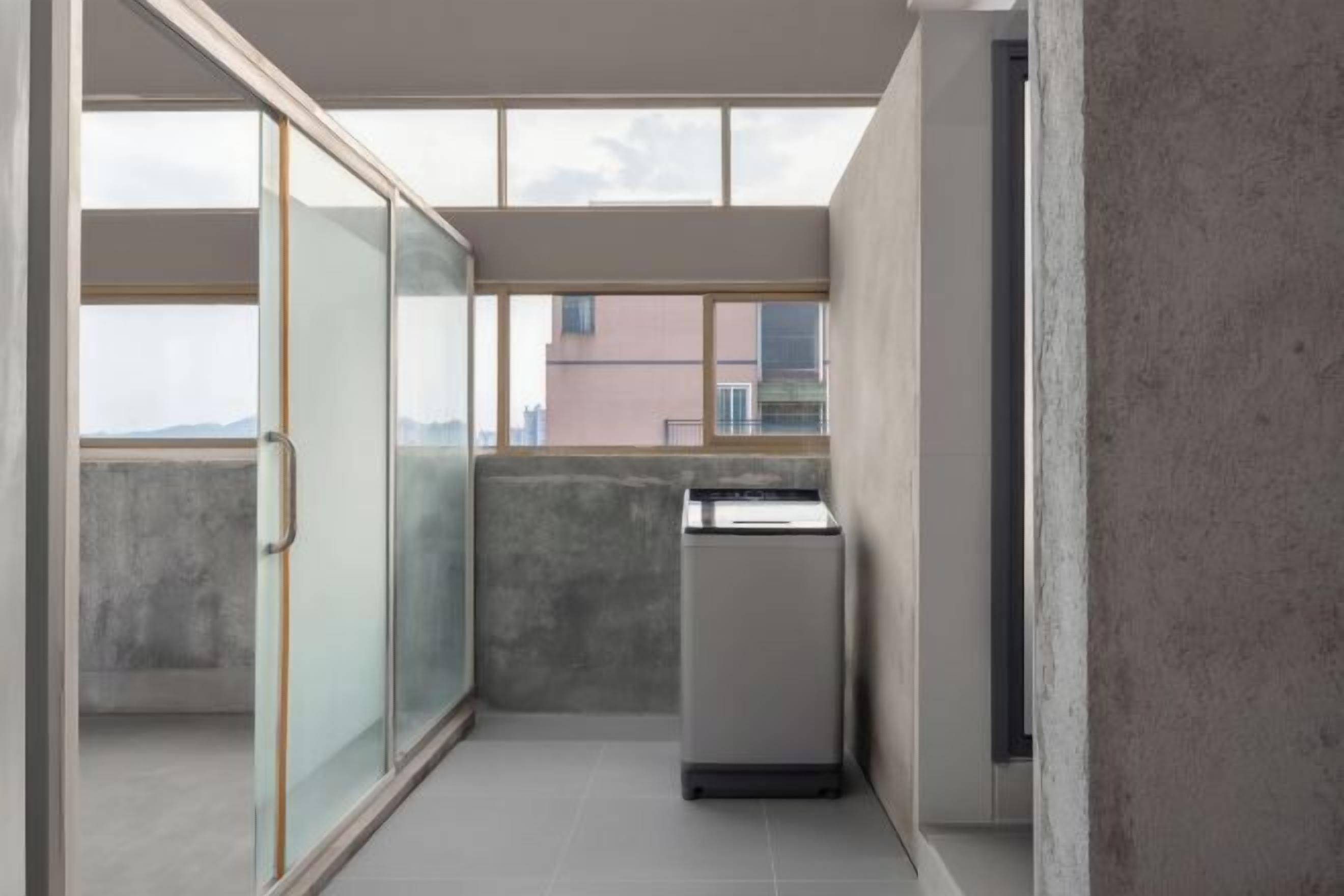 A narrow corridor with a sliding glass door leading to a balcony, a small kitchenette, and a view of buildings in the distance.