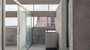 Un couloir étroit avec une porte-fenêtre coulissante donnant sur un balcon, une petite kitchenette et une vue sur des bâtiments au loin.