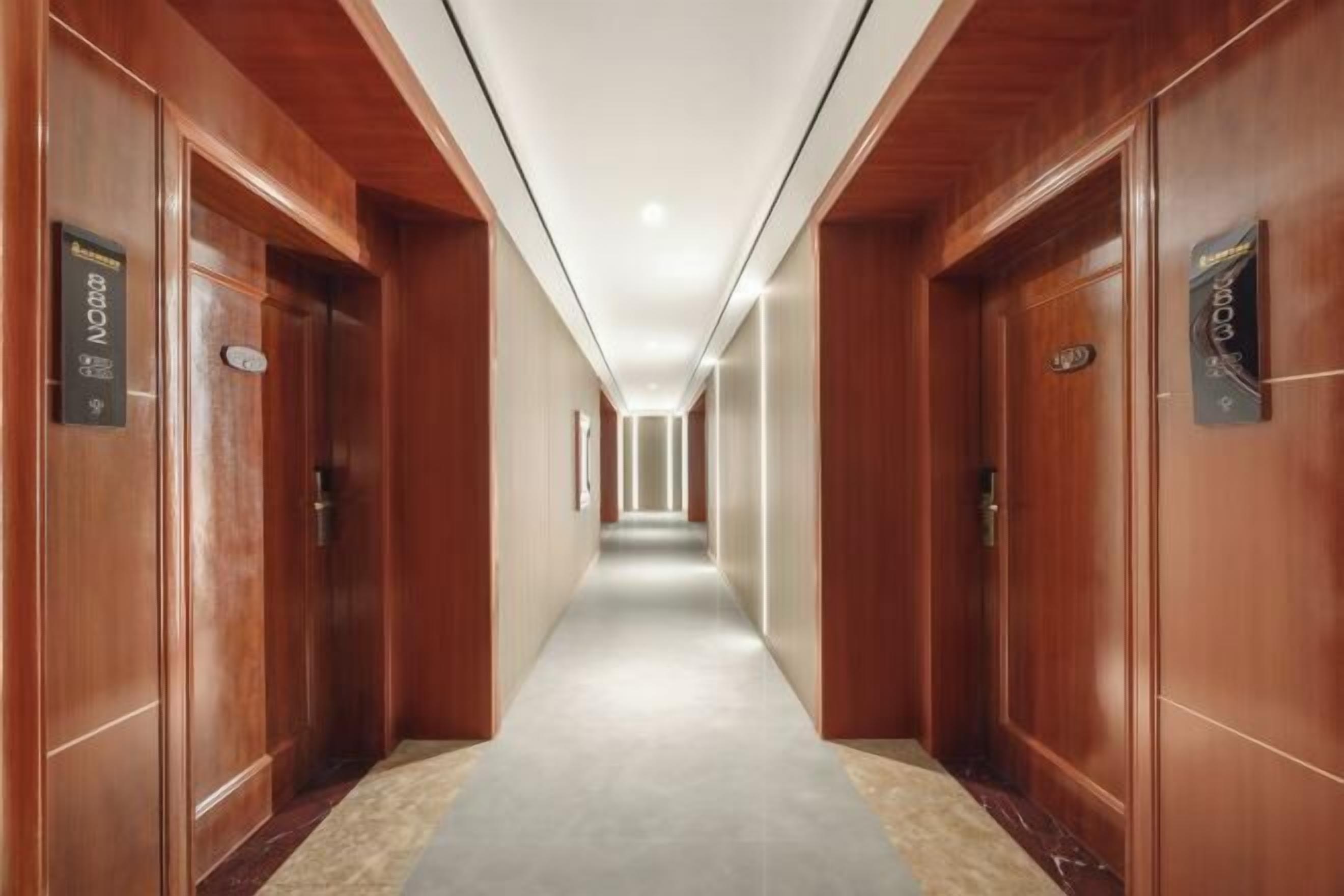 Un corridoio d'albergo con porte in legno, pavimento rivestito di moquette e soffitto con illuminazione a incasso.