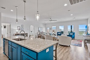 Una cocina amplia con encimera de granito, armarios azules y una isla grande. Hay luces colgantes, un ventilador de techo y vistas al exterior a través de amplias ventanas.